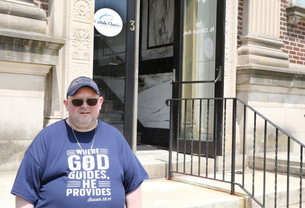 A man in a blue shirt that reads 'Where God guides, he provides' and a blue baseball cap is standing in front of a set of stairs. On the door behind him is a sign for Catholic Charities.