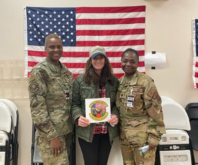 Three women, all smiling, stand facing the camera. An American flag hands in the background.