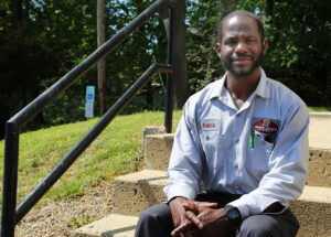 An African American man wearing a uniform shirt is  seated on a set of stairs outside.