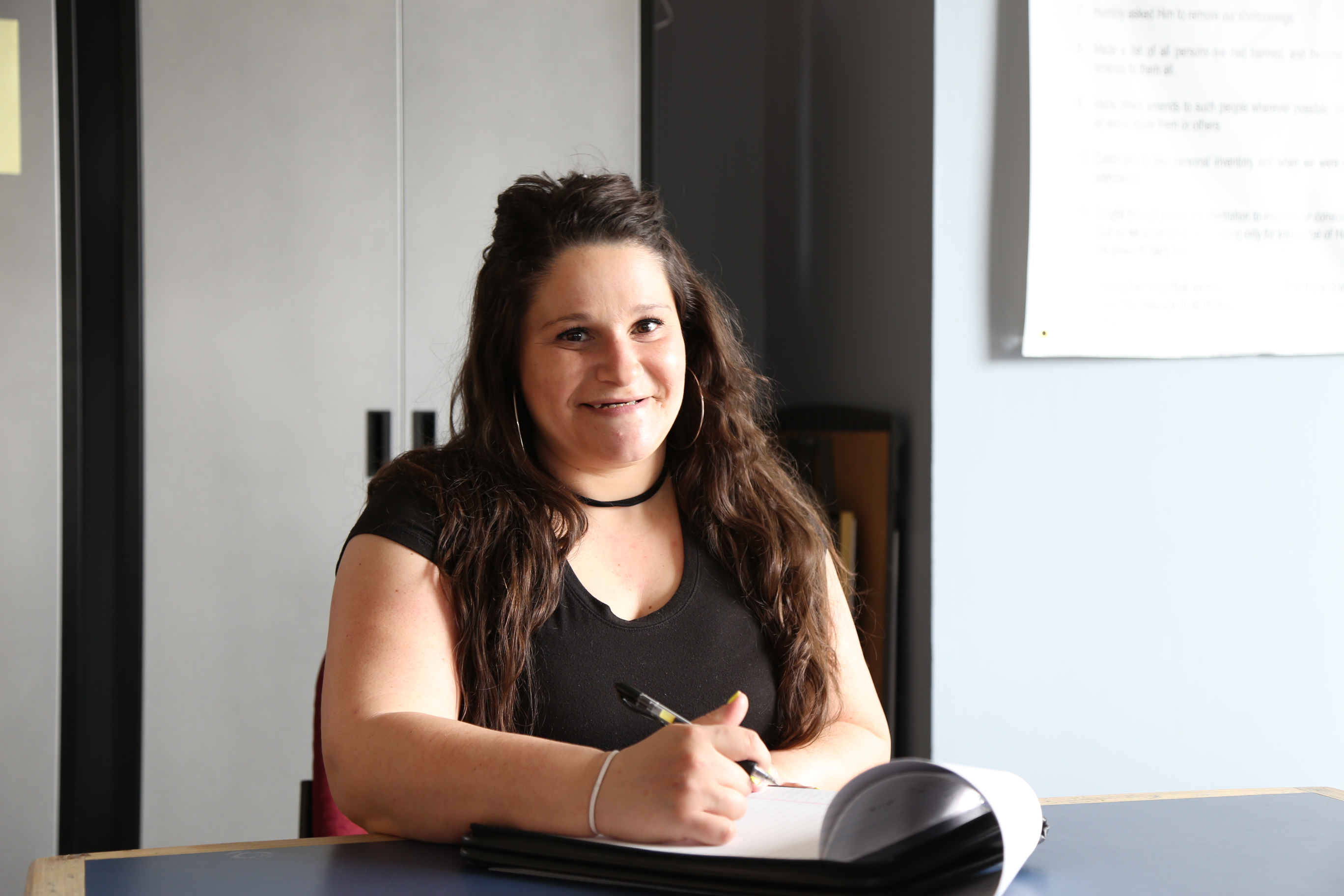 A young woman with brown hair is seated in a classroom holding a pen, poised to take notes. She is smiling