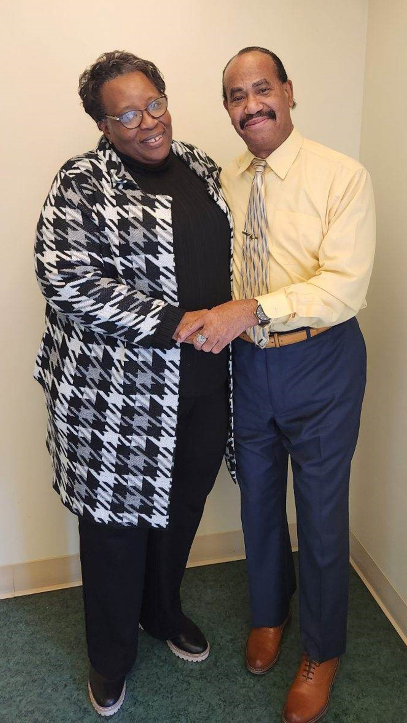An African American woman in a black and white checked jacket and black slacks stands holding hands with an African American Man in a yellow shirt and yellow patterned tie. They are looking at the camera and smiling.