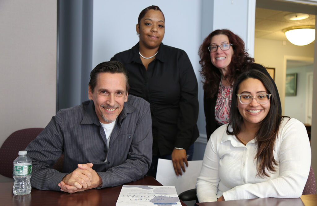 A man and a woman are seated at a table. Two women are standing directly behind them. There are all facing the camera and smiling.