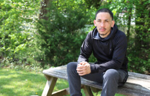 A young man with short black hair wearing a navy blue hoodie is seated on a picnic table. He is looking intently at the camera.