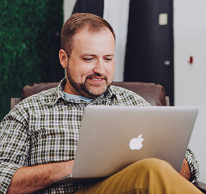 A white man in a  plaid shirt seated looking at a laptop.