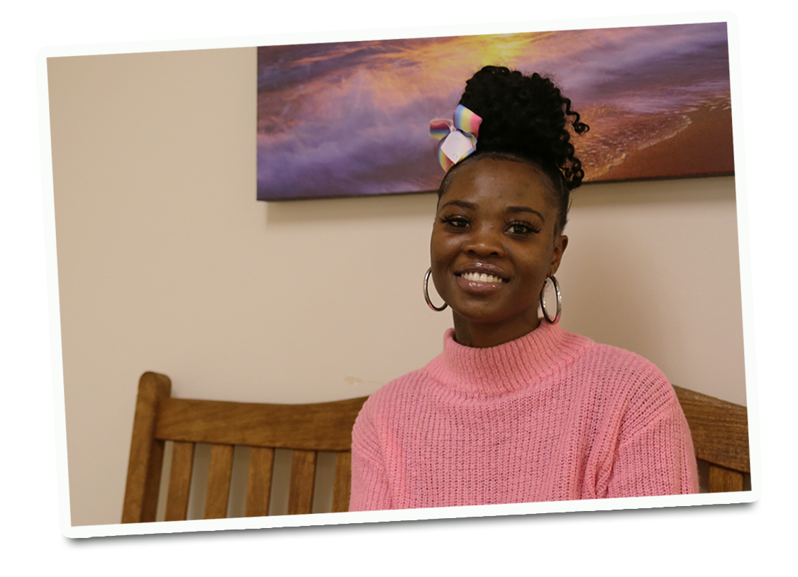 A smiling young, black woman with her hair in a bun on the top of her head wearing a pink turtleneck sweater. 